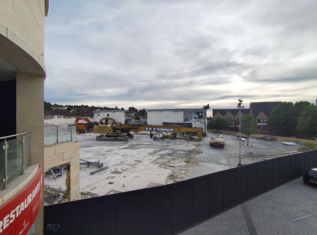 View of the demolition site at Swindon Regent Circus with yellow machinery against a cloudy sky, taken from an upper-level walkway.